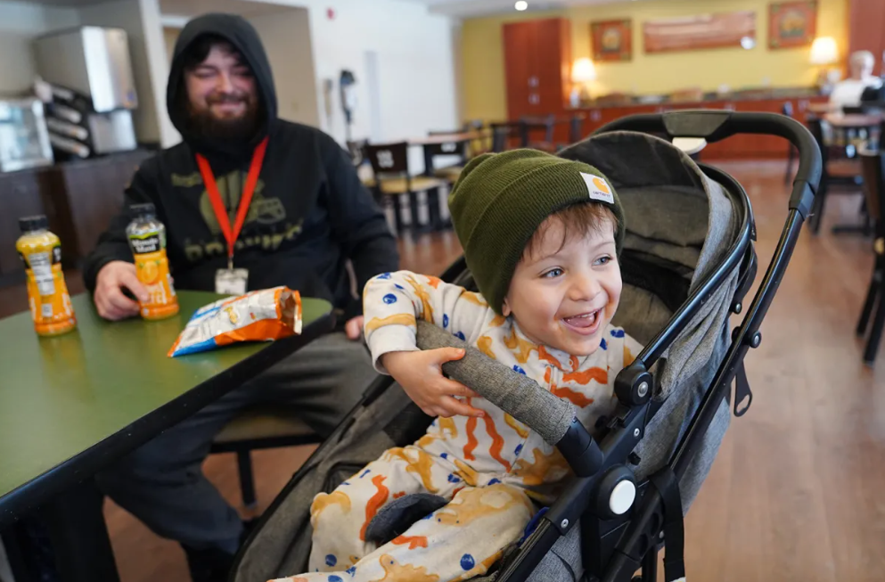 A laughing toddler in a stroller in the House dining room, with an adult seated at the table behind