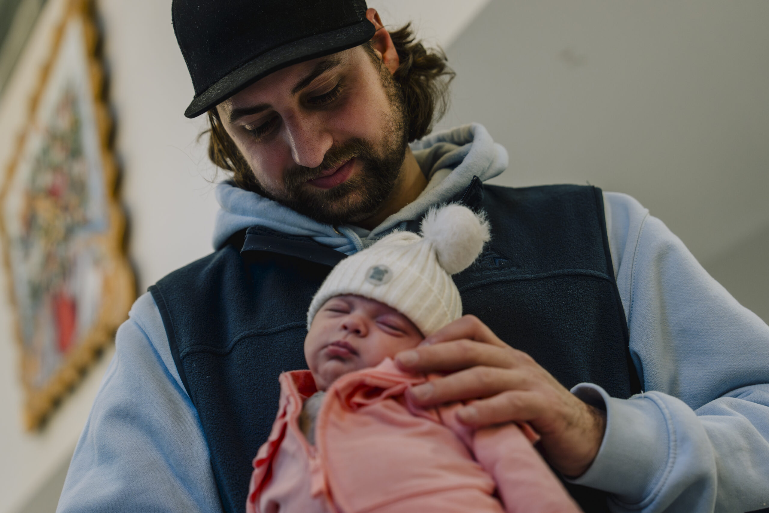 A man in a ball cap looks down at the newborn he is holding
