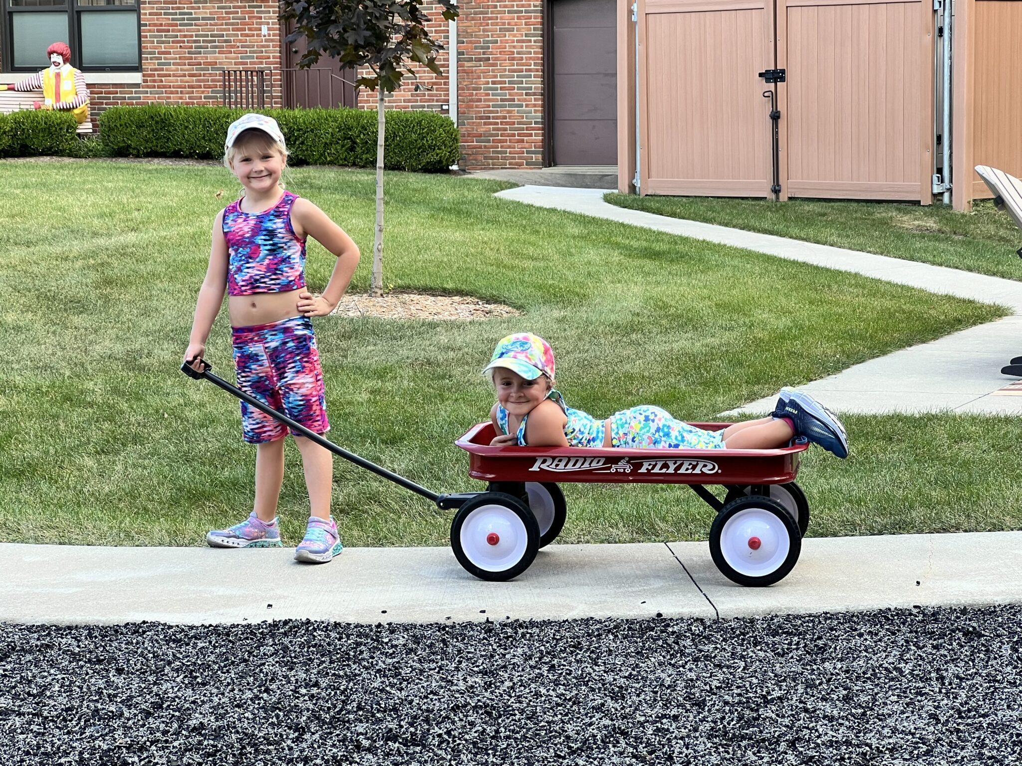 Girls posing for a picture on a sidewalk with a radio flyer red wagon one girl is laying down in the wagon turned toward the camera smiling the other girl is holding the handle And grinning with one hand on her hip