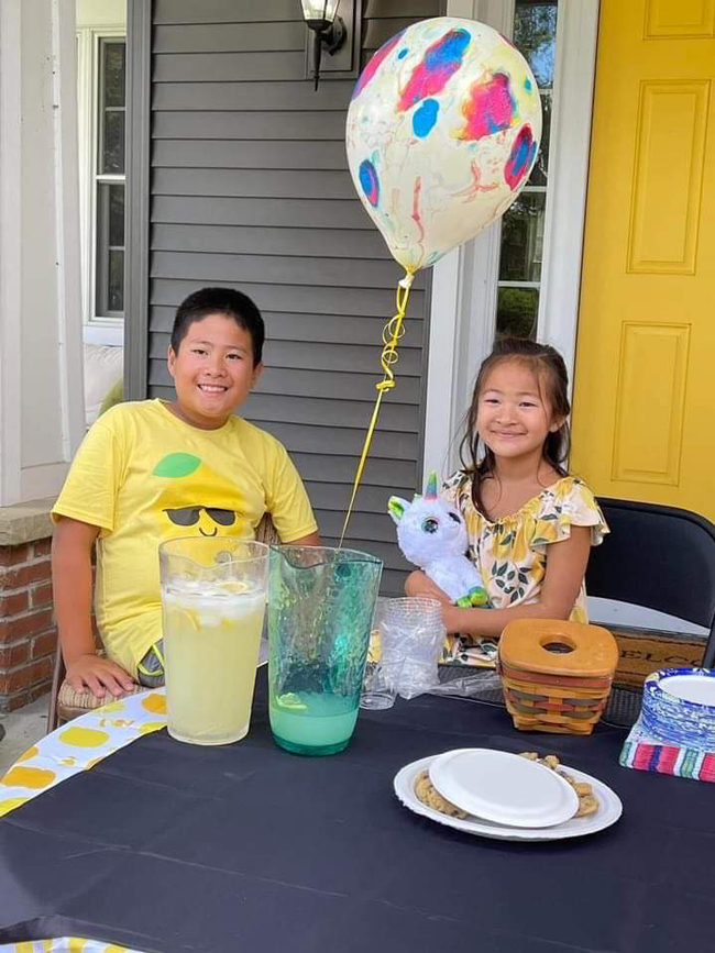 Two children run a lemonade stand fundraiser, with a balloon, cookies, and pitchers of lemonade on the table