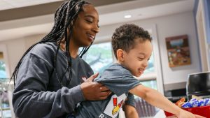 A woman smiles as she holds a toddler who is reaching toward a table
