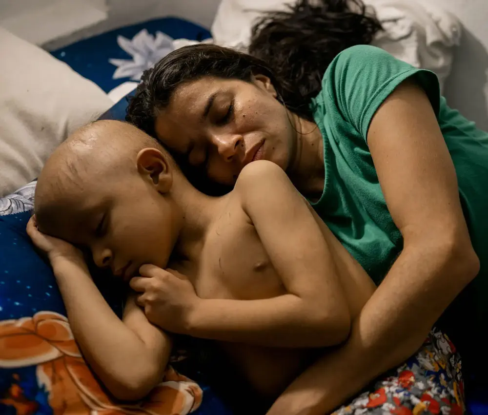 A mother lies beside her young child, holding him close as he sleeps