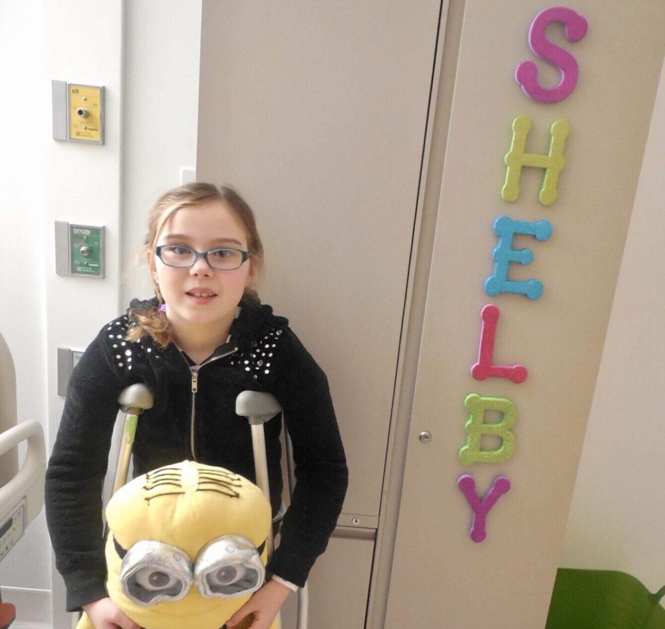 little girl on crutches holding minion stuffed animal. her name, Shelby, is spelled out in colorful letters on the wall behind her
