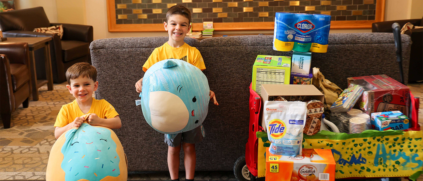 Two young boys hold plush toys beside a cart stacked with donated household supplies in a House lobby