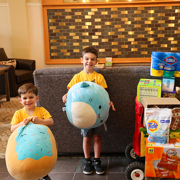 Two young boys hold plush toys beside a cart stacked with donated household supplies in a House lobby
