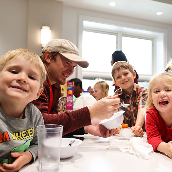 An adult serves cereal to three laughing children at a kitchen counter
