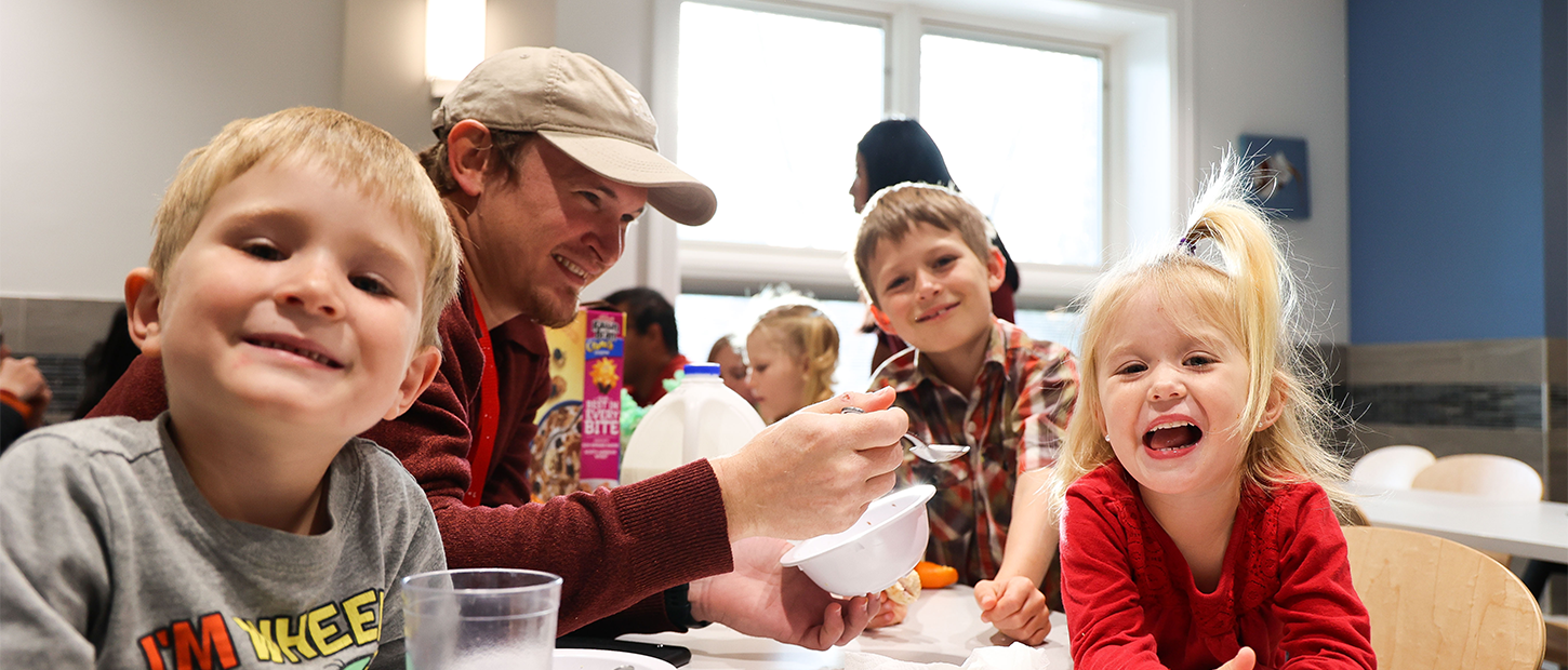 An adult serves cereal to three laughing children at a kitchen counter