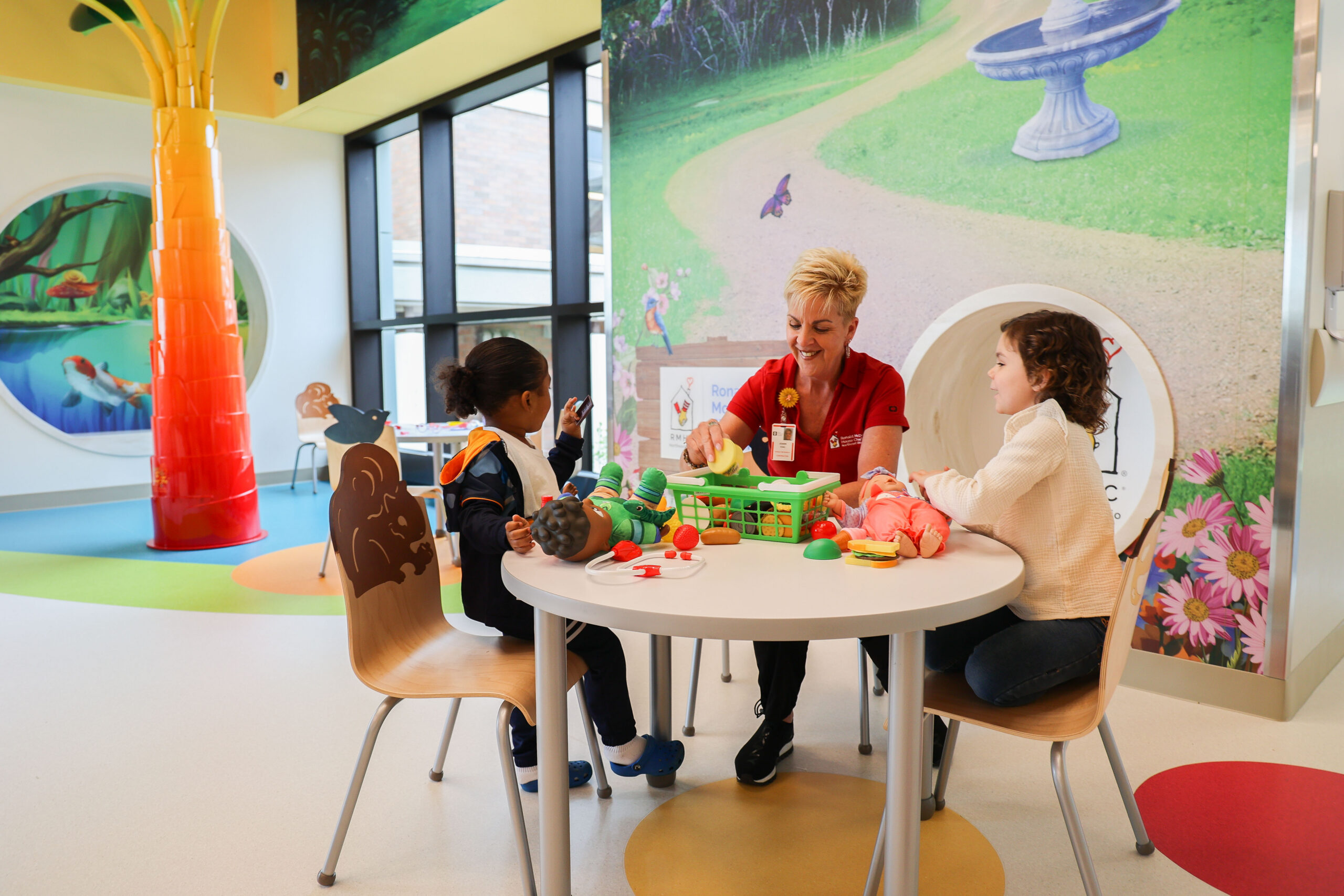 A staff member plays with two children at a table in a brightly decorated playroom