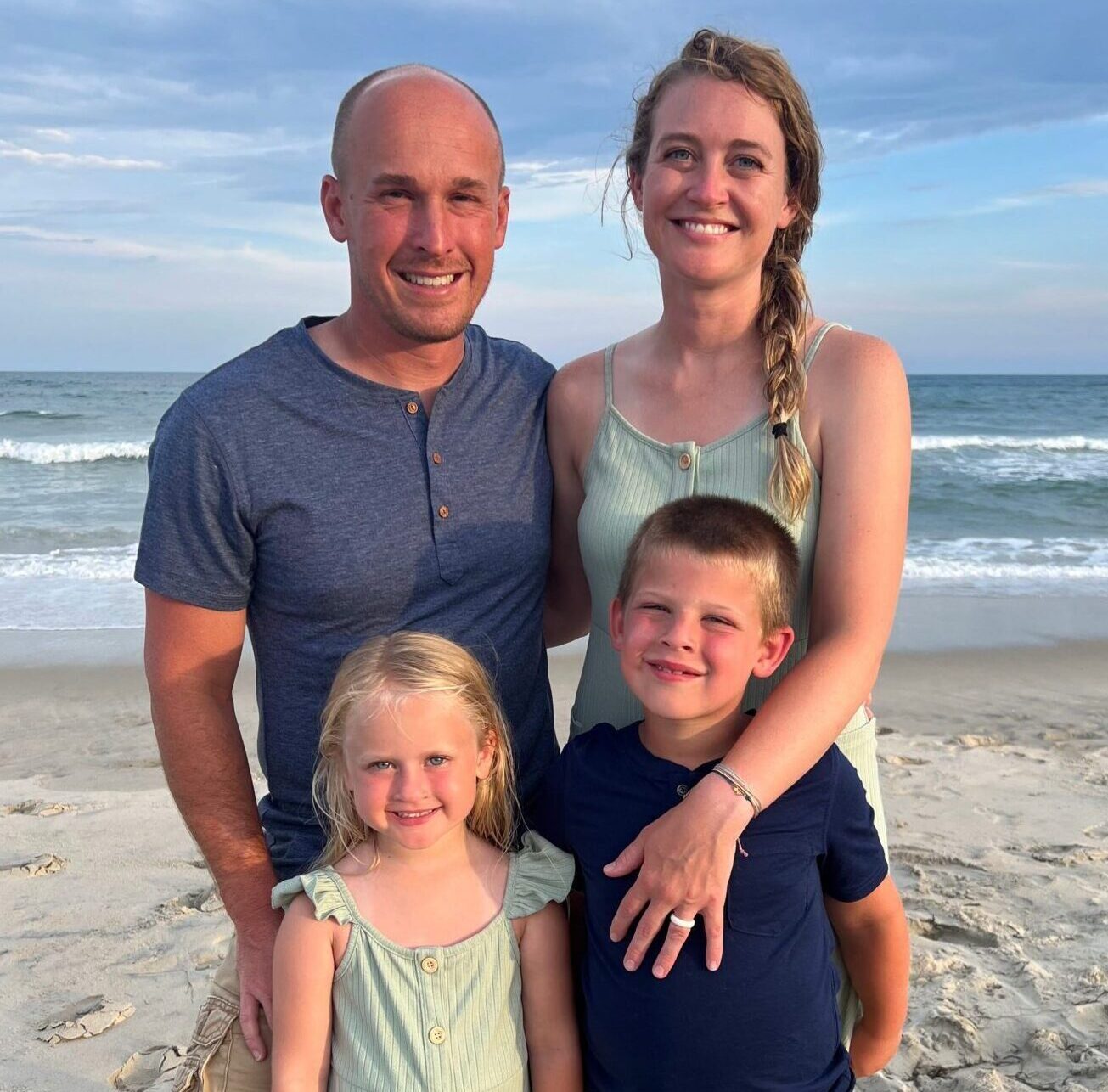 The Brown family standing on the beach. Mom, Dad, Sister and brother smile and pose with their back to the water