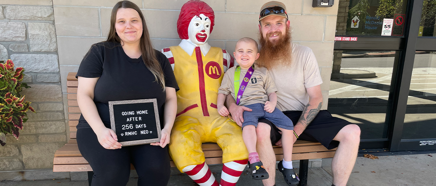 A family sits with a Ronald McDonald statue holding a sign that reads 'Going home after 256 days, RMHC NEO'
