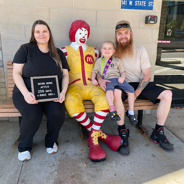 A family sits with a Ronald McDonald statue holding a sign that reads 'Going home after 256 days, RMHC NEO'