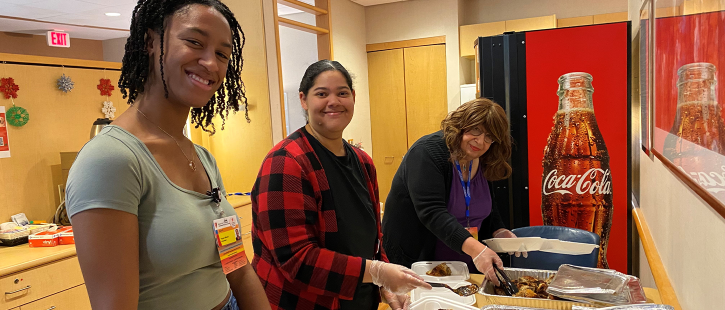 Three volunteers serve a meal from foil trays in a Family Room kitchen