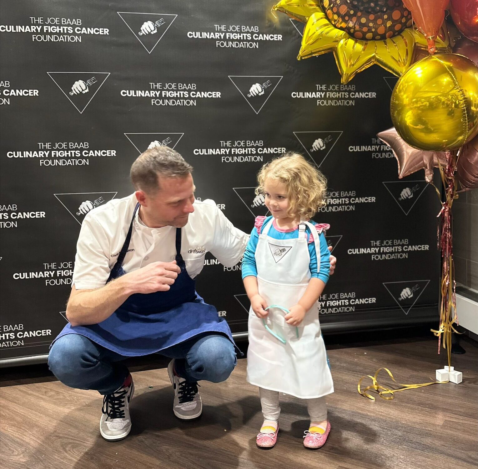 A chef crouches down next to a little girl also wearing a kitchen apron. They are posing in front of a banner that says "The Joe Baab Culinary Fights Cancer Foundation". There is a bunch of bright gold balloons shaped like sunflowers and stars floating up from the floor next to them