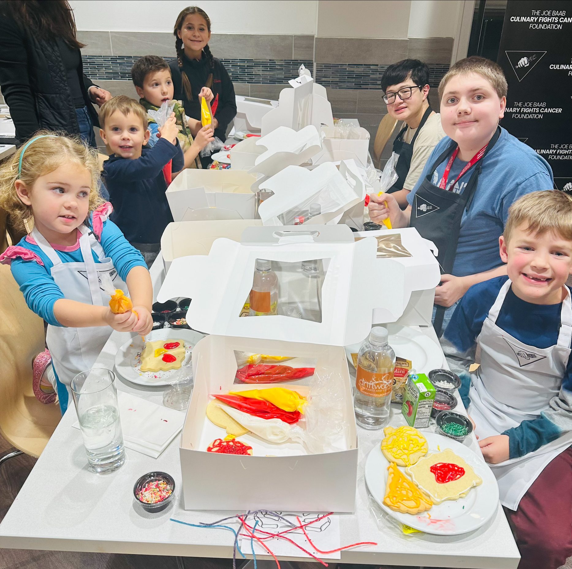Seven kids are gathered around a long table with white boxes and cookie frosting supplies arranged down the middle