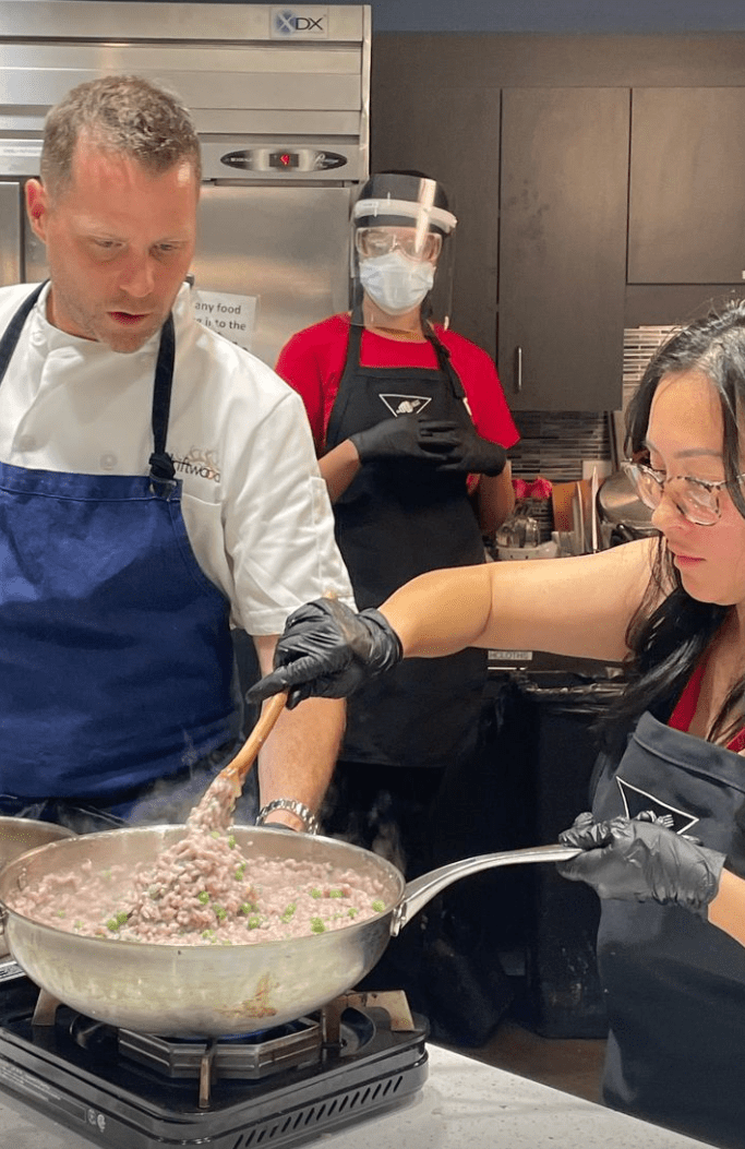 In a commercial kitchen, a young woman is stirring a big wok full of food on a burner. There is a man in a blue apron looking on and another person in a black apron behind them also watching.