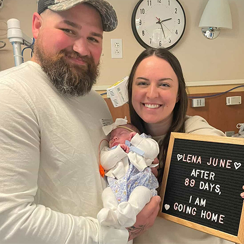 Parents hold their newborn in a hospital room beside a sign that reads 'Lena June, after 89 days, I am going home'