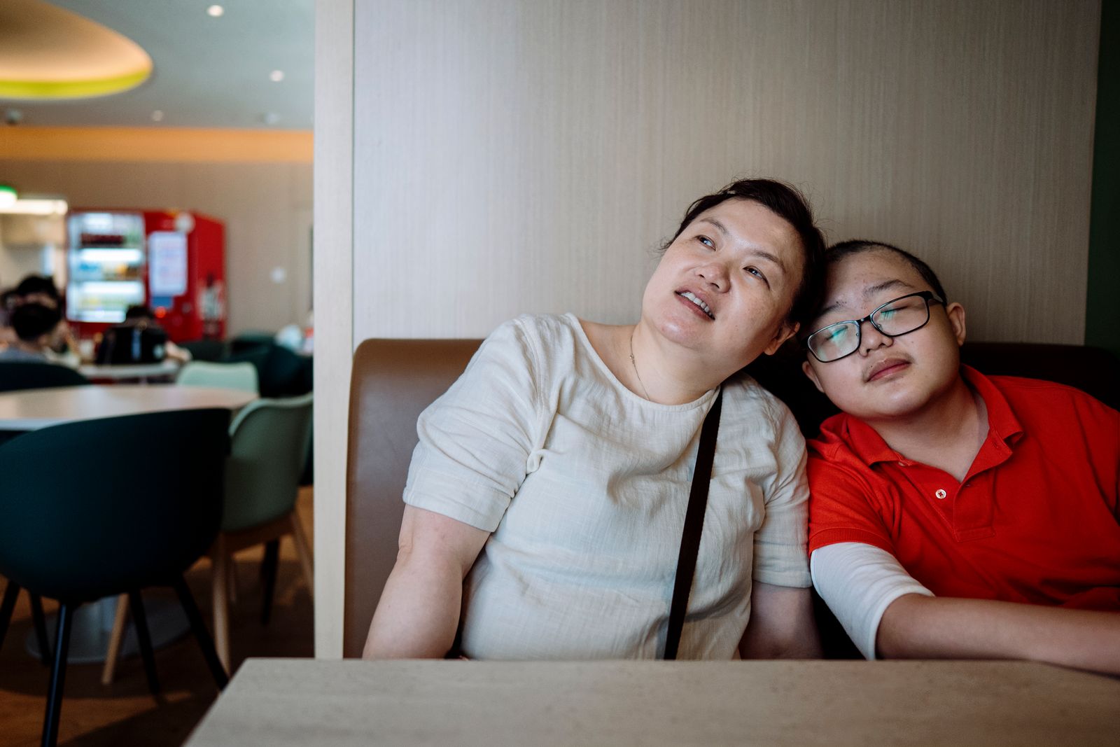 A woman and a teenage boy rest their heads together in a restaurant booth