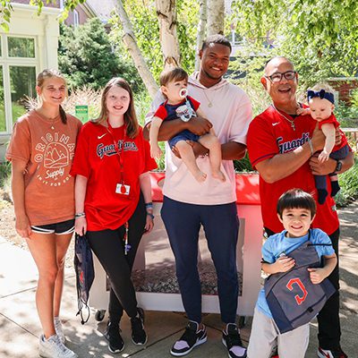 Cleveland Guardians player Oscar Gonzalez standing in the yard of the Cleveland Ronald McDonald House wiht two teenage girls, a dad and three yound boys. Oscar is holding a baby.