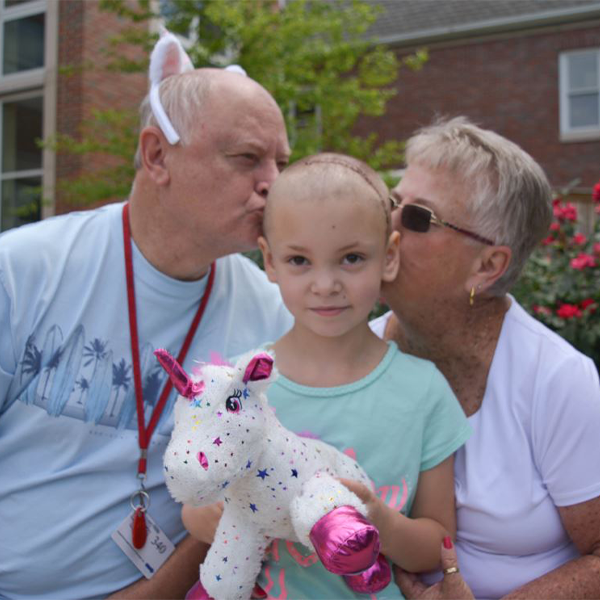 An older man and woman kiss the cheeks of a young girl who is holding a unicorn stuffed animal